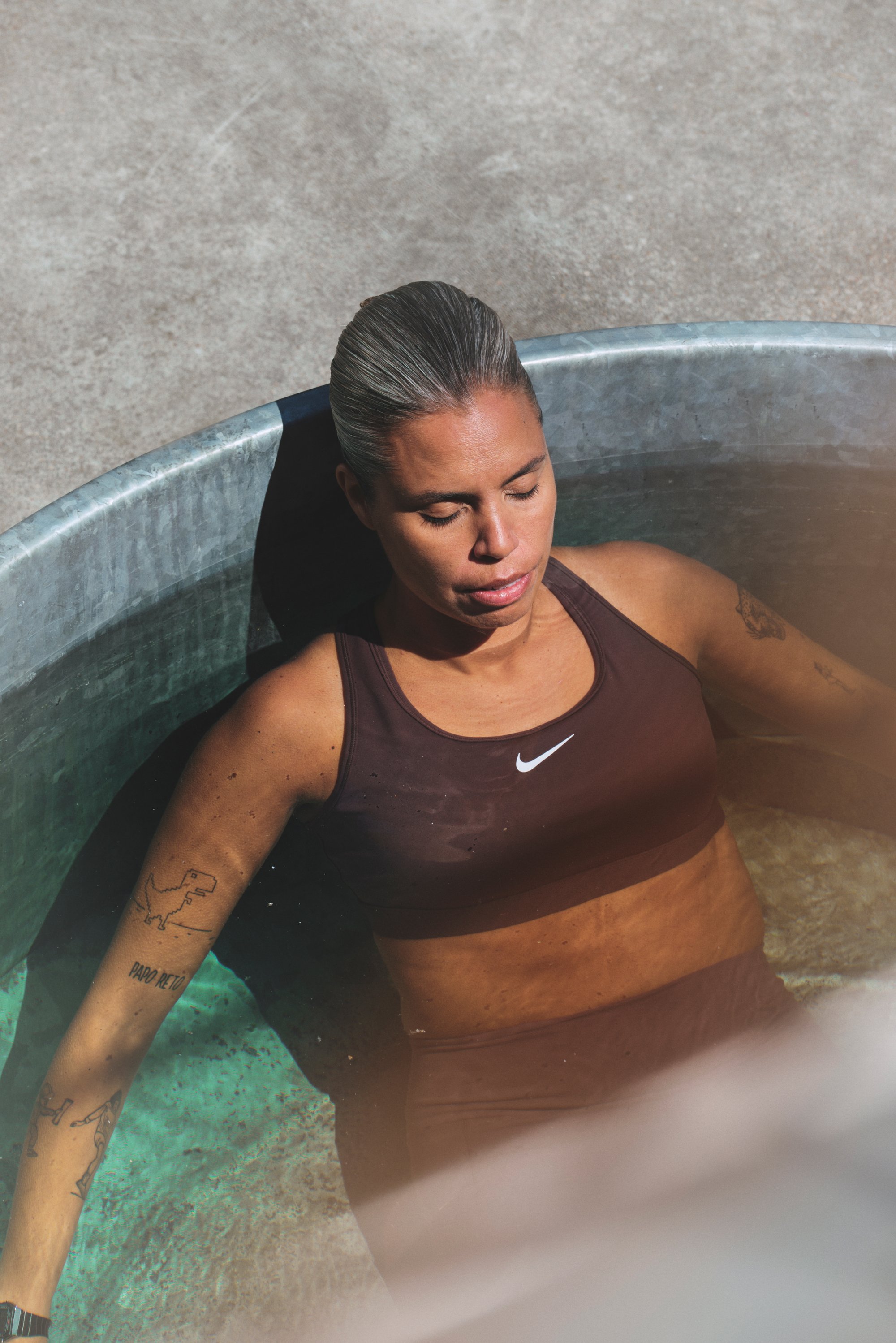 Woman with slicked-back hair relaxes in a metal tub filled with water, wearing a brown sports bra. Sunlight reflects off the water’s surface.