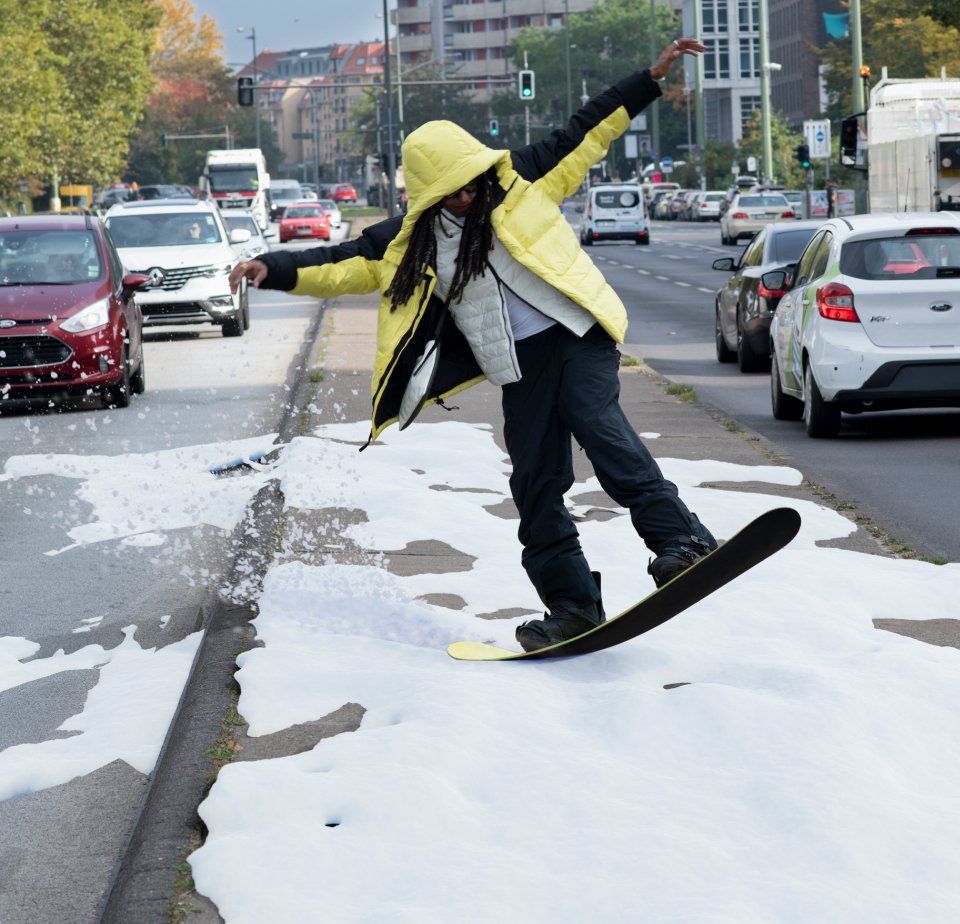 A person in a yellow jacket snowboards on a city street, surrounded by cars and a flowing spray of snow. Urban environment visible.