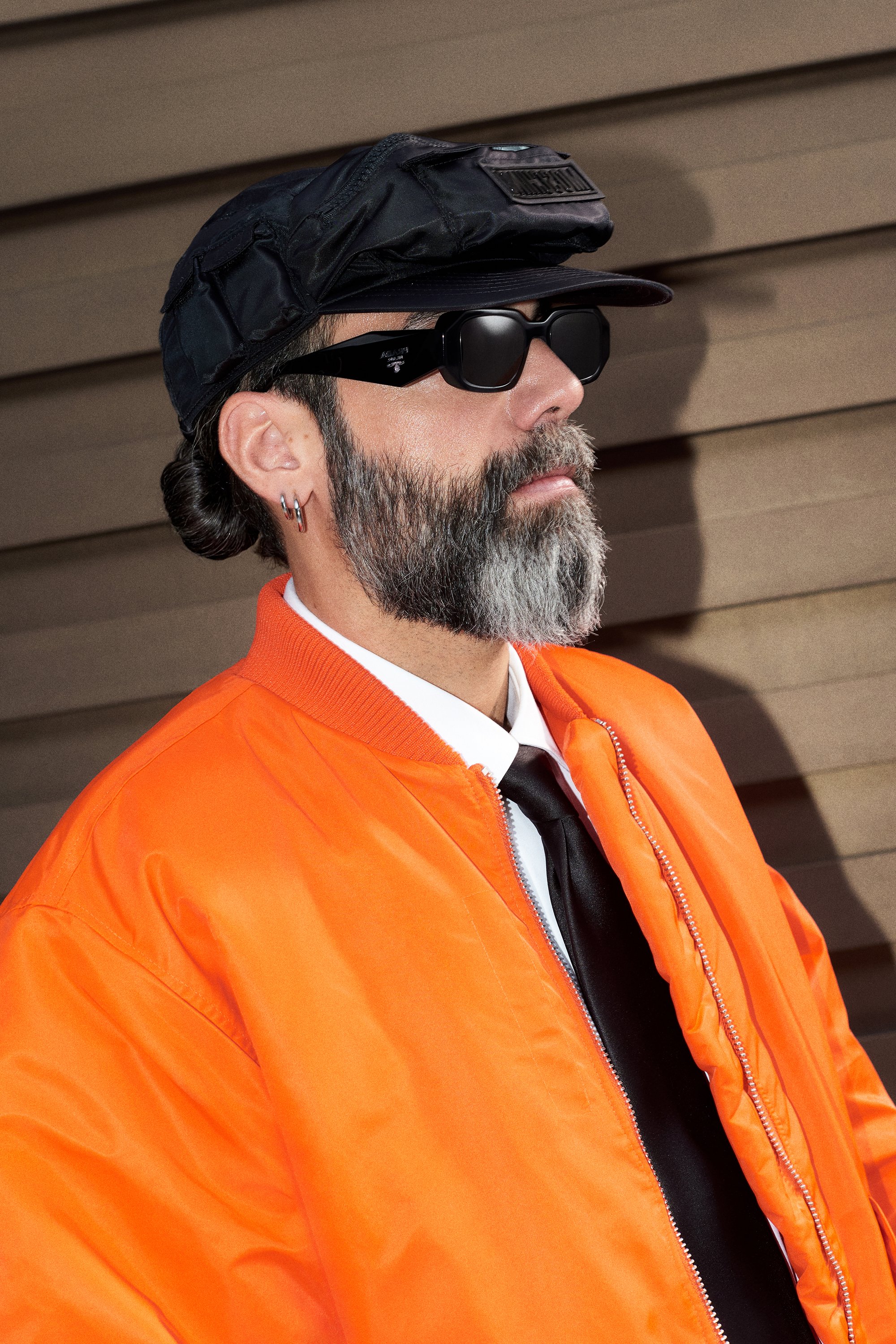A man with a beard wears sunglasses, an orange bomber jacket, and a black cap, posing against a textured brown wall.