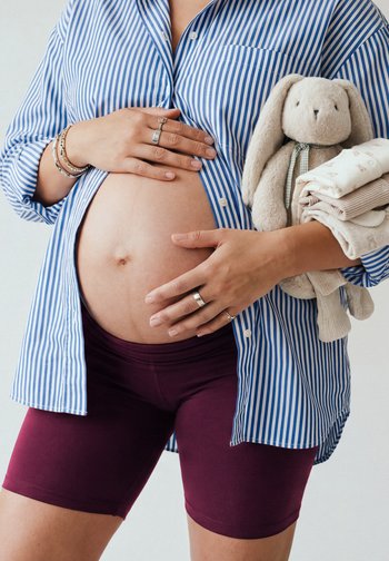 Nosečnica v modri srajci z belimi črtami in bordo kratkih hlačah z eno roko nežno drži trebuh, v drugi pa plišastega zajčka in otroška oblačila.