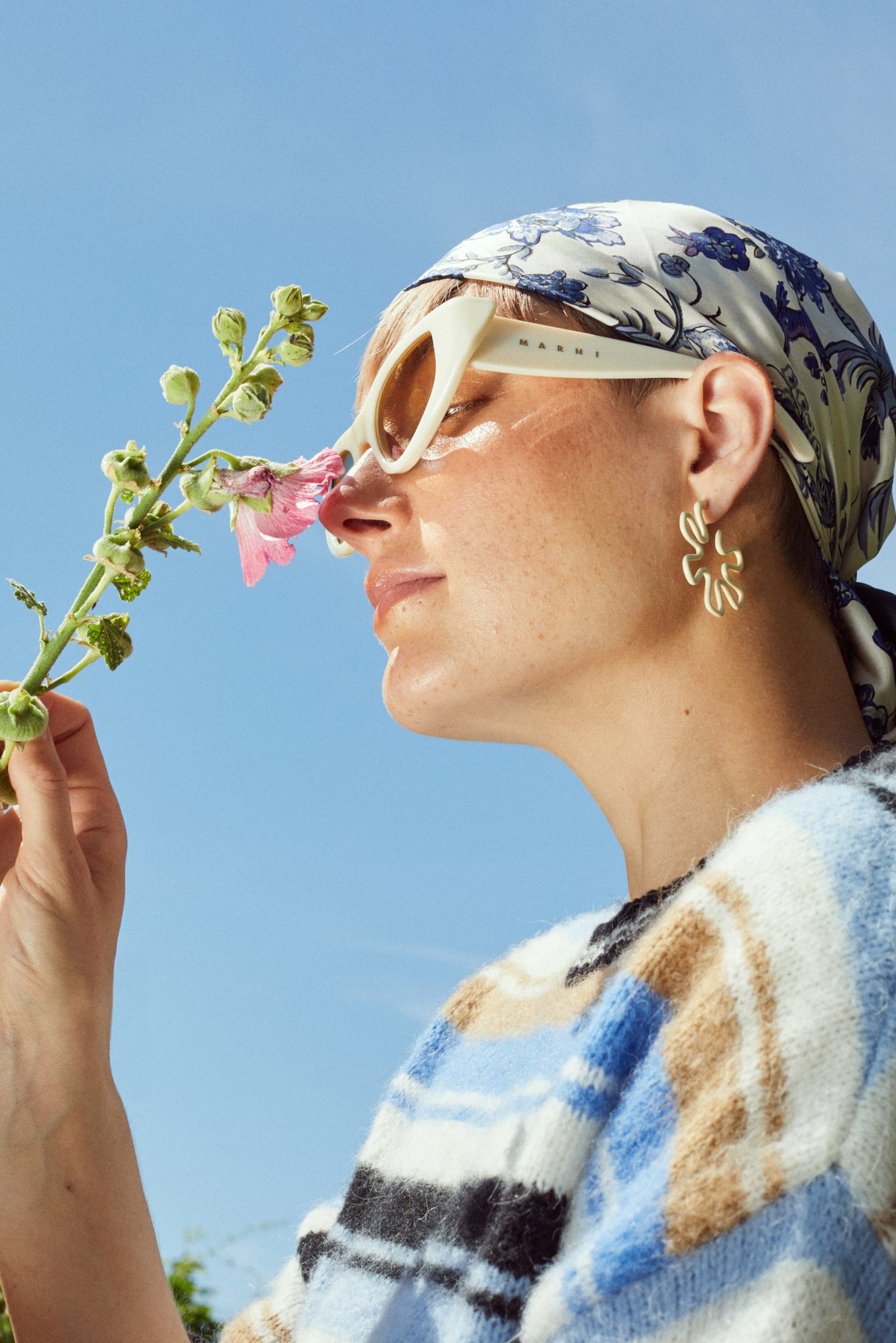 Una mujer con gafas de sol y una bufanda floral sostiene suavemente una flor cerca de su cara, rodeada de un cielo azul claro. Ella lleva un suéter estampado.