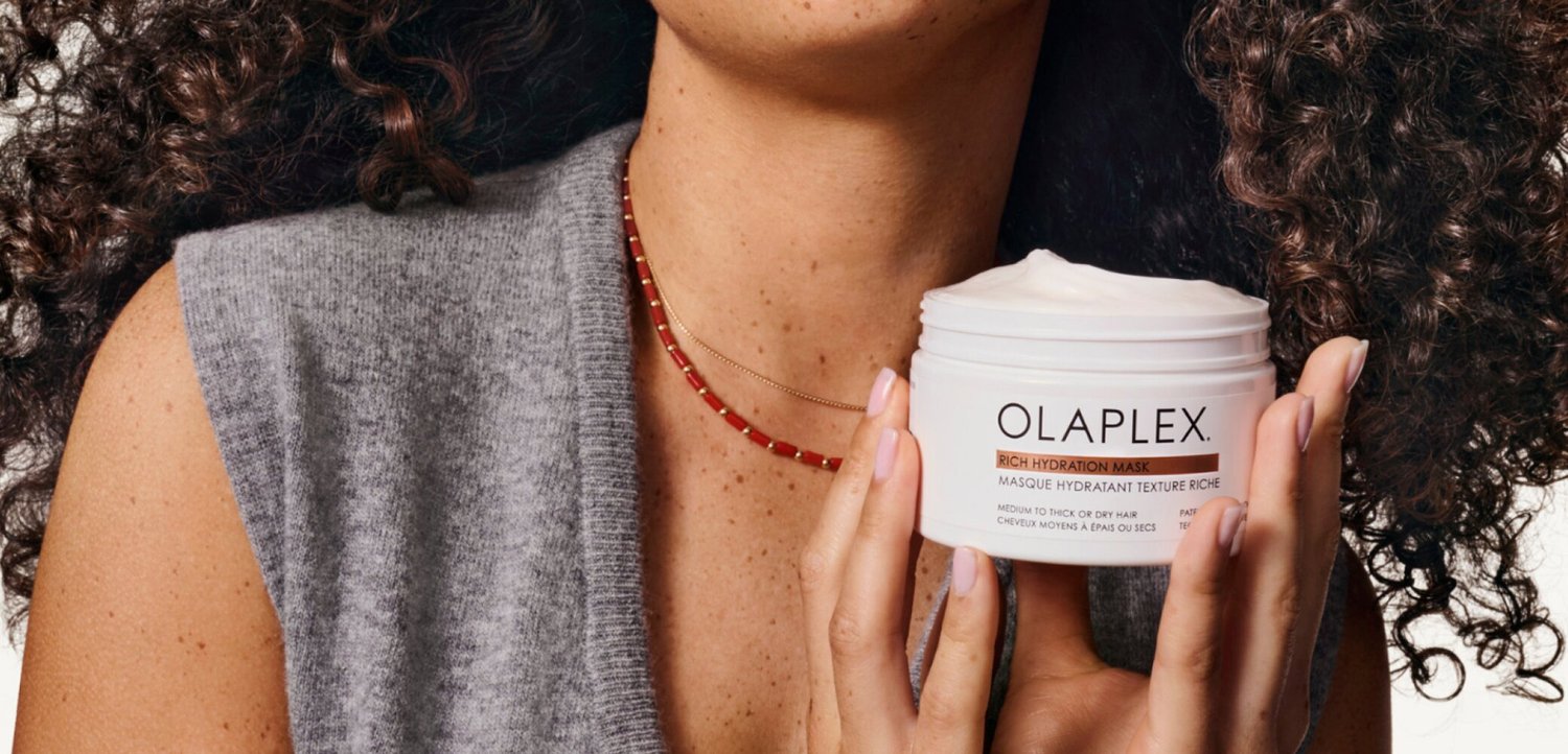 Persoon met krullend haar die een witte pot vasthoudt met het etiket Olaplex Rich Hydration Mask bij hun nek en borst.