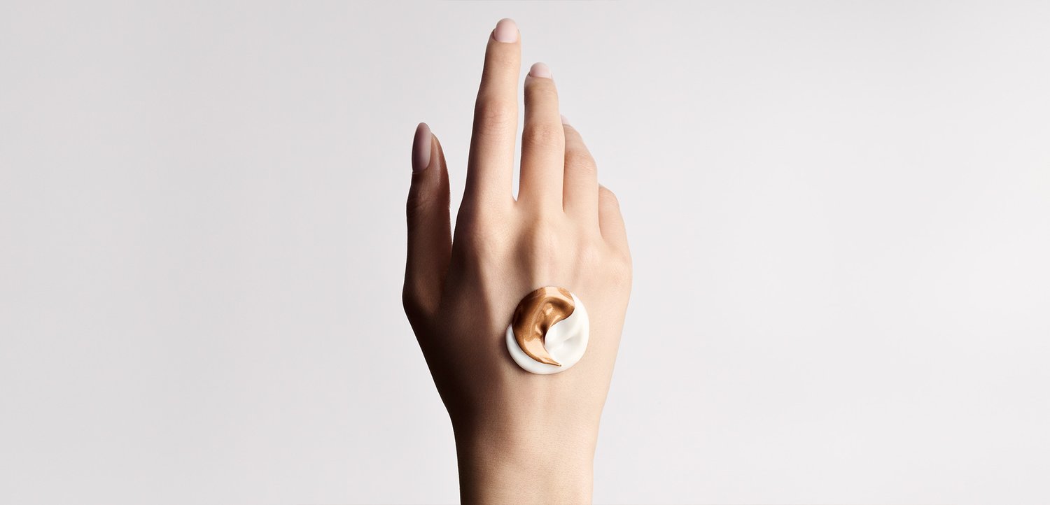 A hand with a dollop of two-tone cream, white and bronze, on the back against a plain light background.