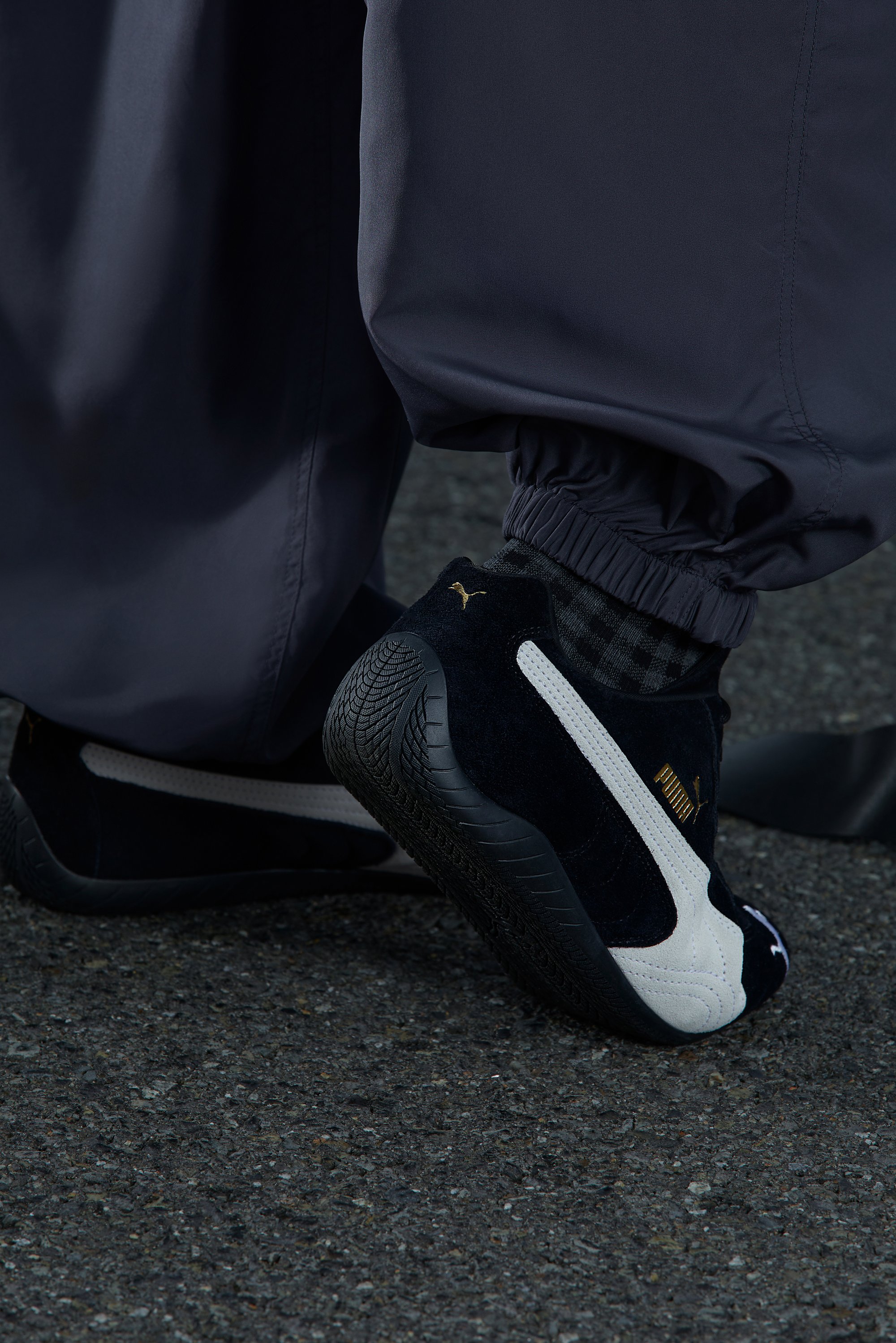Zwart-witte Puma sneakers met een gouden logo, gedeeltelijk verborgen onder loszittende donkergrijze broek, tegen een gestructureerd asfaltoppervlak.