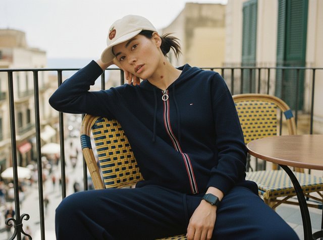 Jeune personne en survêtement bleu marine et casquette blanche assise de manière détendue sur le balcon d’un café en extérieur avec un paysage urbain en arrière-plan.