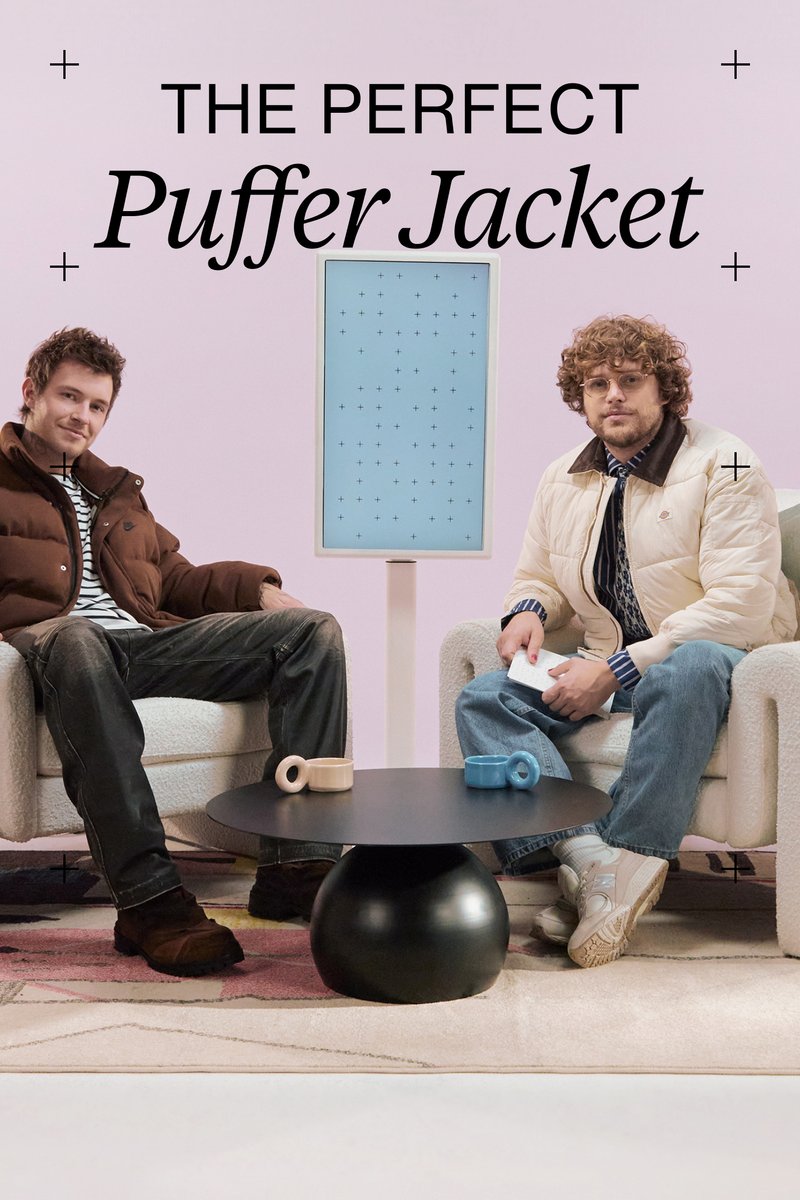 Two men sit on cream sofas in a pastel pink setting. A black table holds colorful mugs while a light blue display stands between them.