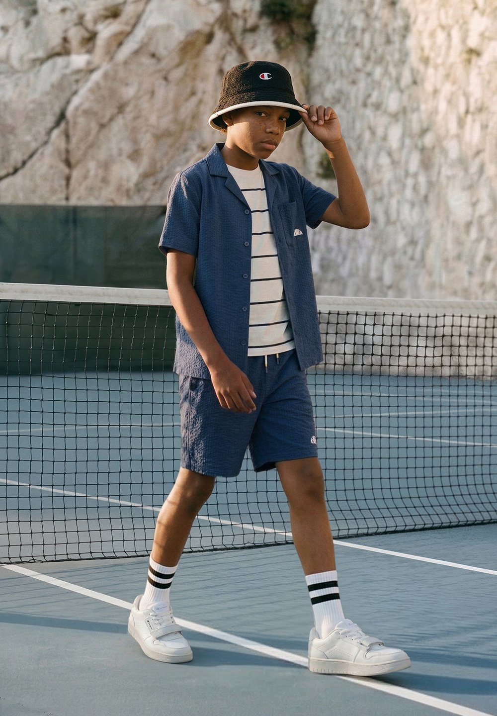 Poika sinisissä shortseissa, laivastonsinisessä paidassa, raidallisessa t-paidassa ja mustassa kalastajahatussa kävelee tenniskentällä lähellä verkkoa.