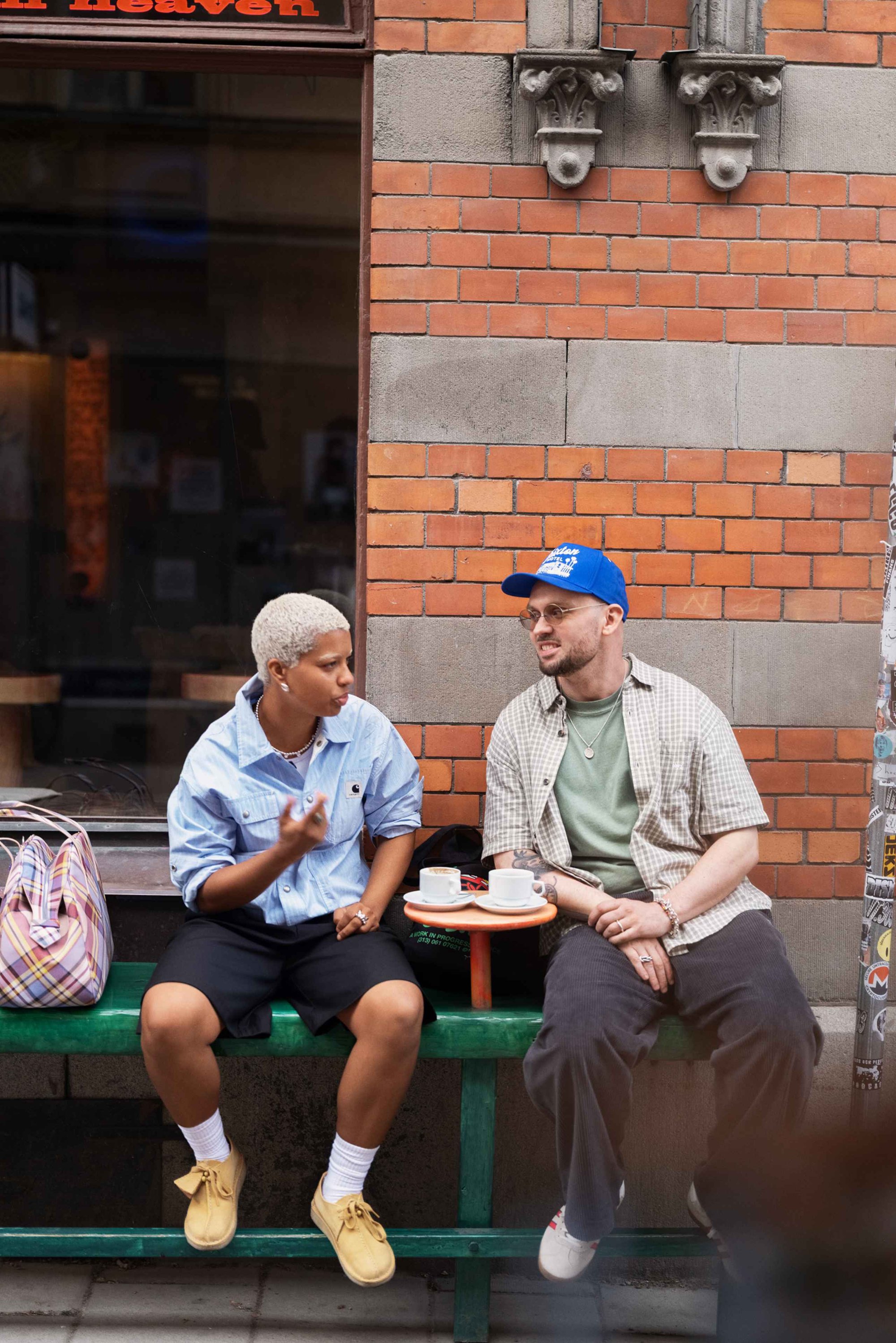 Twee individuen zitten op een groene bank, de een draagt een lichtblauw shirt en zwarte shorts, de ander een geruit shirt, met koffiekoppen op een kleine tafel.