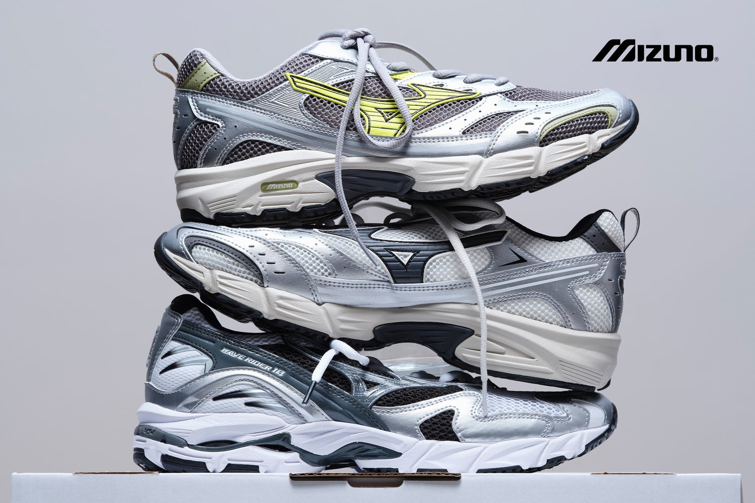 Tres pares de zapatillas de correr Mizuno apiladas verticalmente, con diseños en gris y plata con acentos y cordones neón.