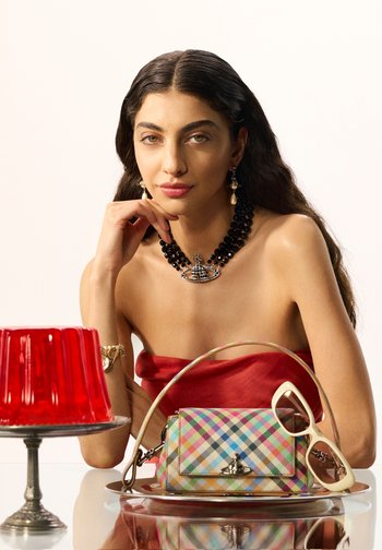 Femme aux longs cheveux foncés portant un haut rouge sans bretelles, un collier noir perlé et des boucles d'oreilles dorées, assise à une table avec un sac à main à carreaux colorés et des lunettes de soleil beige.