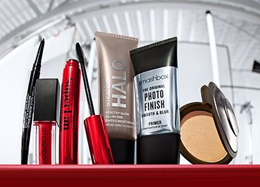 Varios productos de maquillaje dispuestos en una estantería roja, incluyendo máscara de pestañas, brillo de labios, humectante, prebase y bronceador, sobre un fondo neutro.