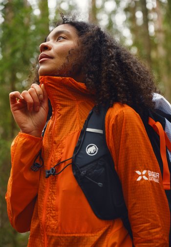 Kvinne med krøllete hår som går tur i skogen, iført oransje jakke og svart ryggsekk, justerer jakkekraven.