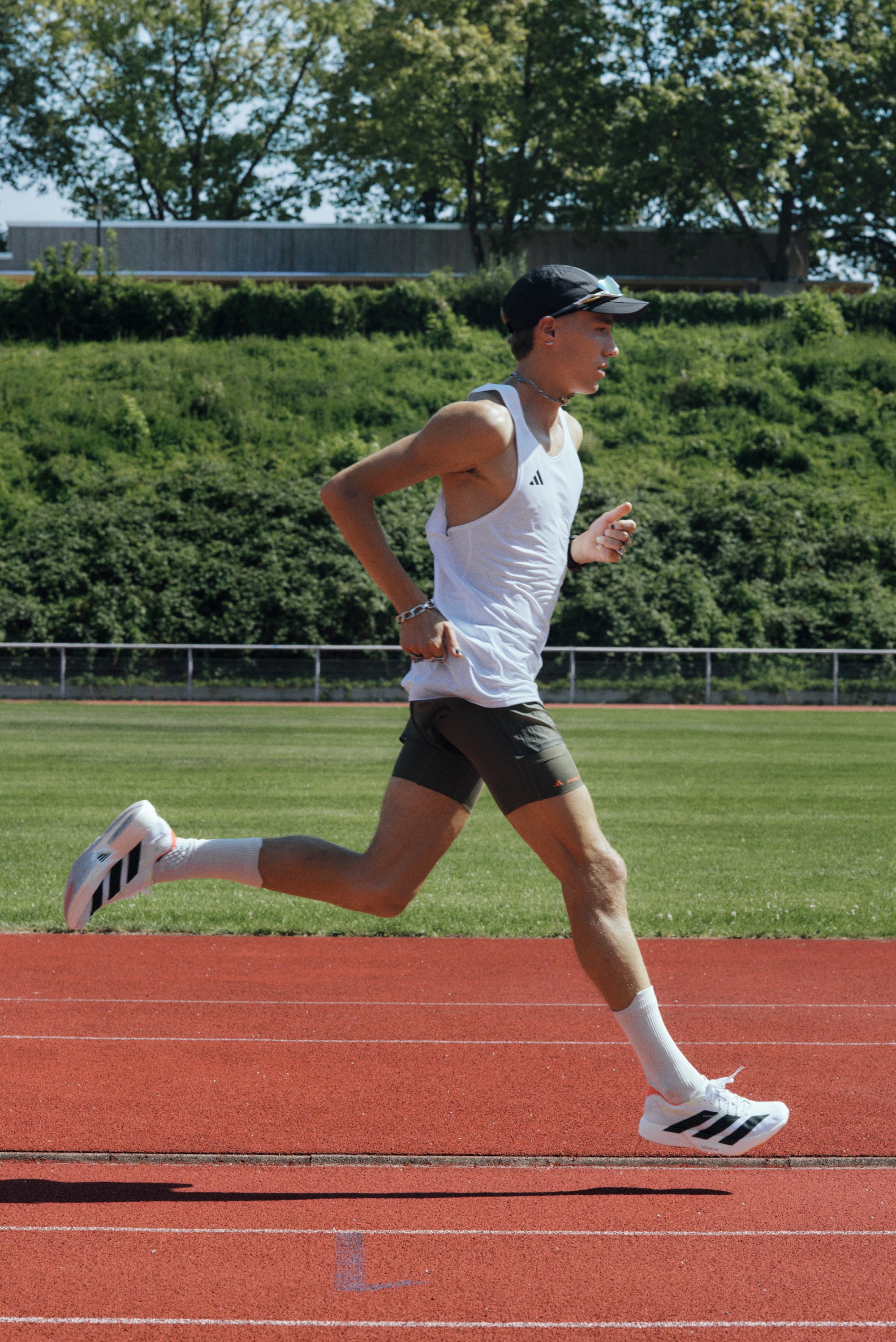Sportieve schoenen met zwarte strepen, witte bovenkant en grijze shorts. Een hardloper in een mouwloos wit shirt en een zwarte pet op een rode baan.