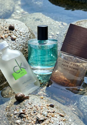 Trois flacons de parfum, dont Calvin Klein CK One et Issey Miyake L'Eau d'Issey Pour Homme, placés sur des rochers mouillés avec de petits escargots.