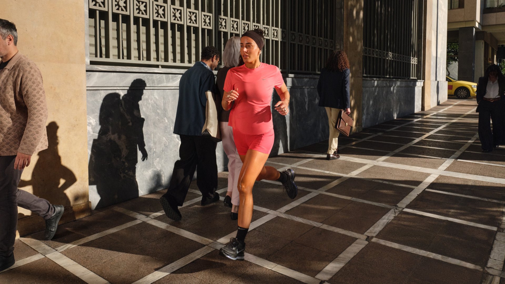 Femme portant des vêtements de sport roses en train de courir sur un trottoir en ville, tandis que plusieurs piétons marchent à proximité en plein jour.