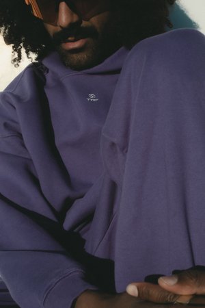 Lila Sweatshirt mit lockerer Passform, ausgestattet mit einem kleinen weißen Logo. Glatte Textur, gerippte Bündchen und ein Akzent von orangefarbenen Sonnenbrillen.