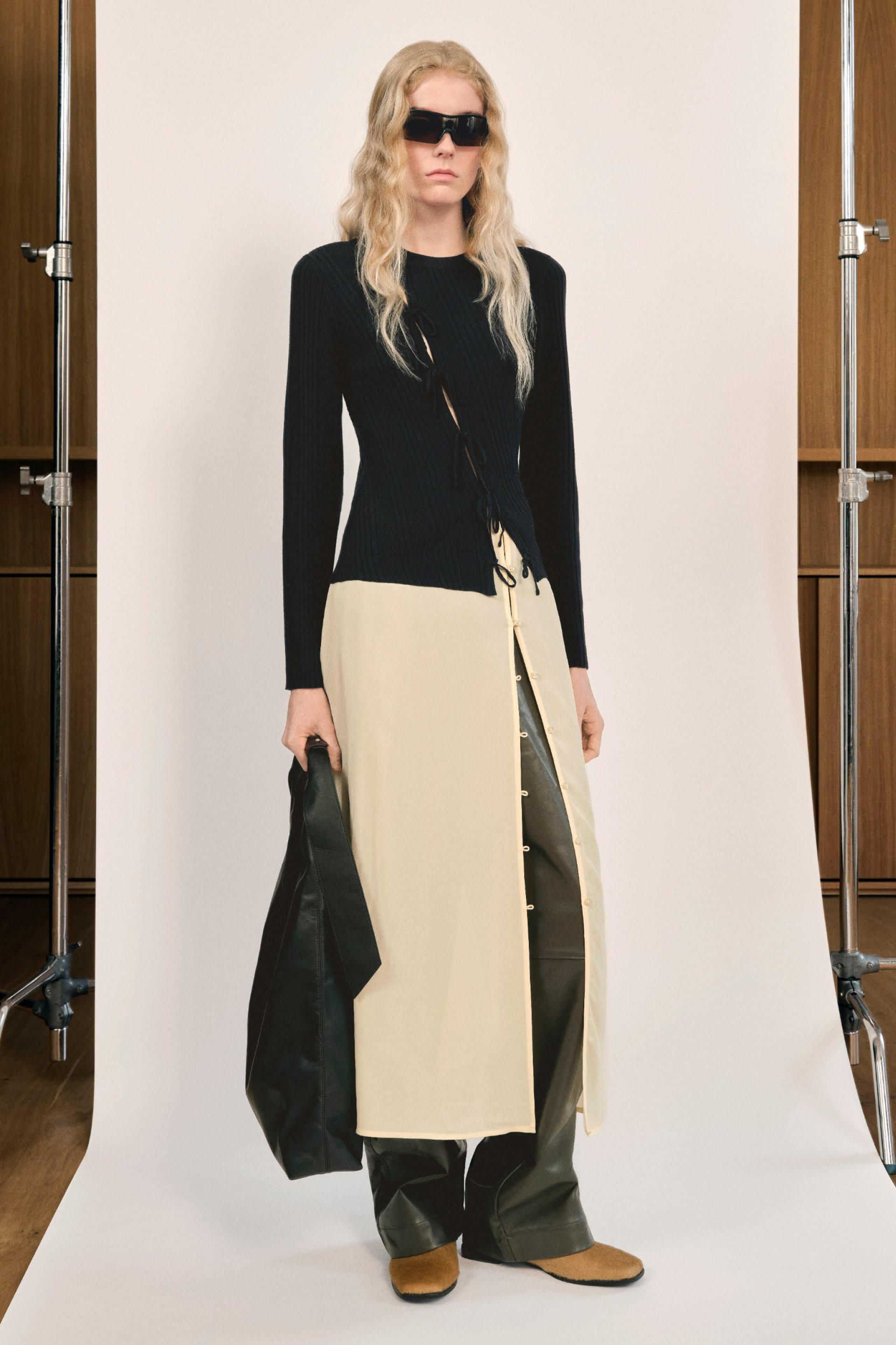Una modelo con cabello largo y ondulado lleva un suéter negro acanalado y un vestido de color crema, sosteniendo un bolso negro frente a un fondo neutro.