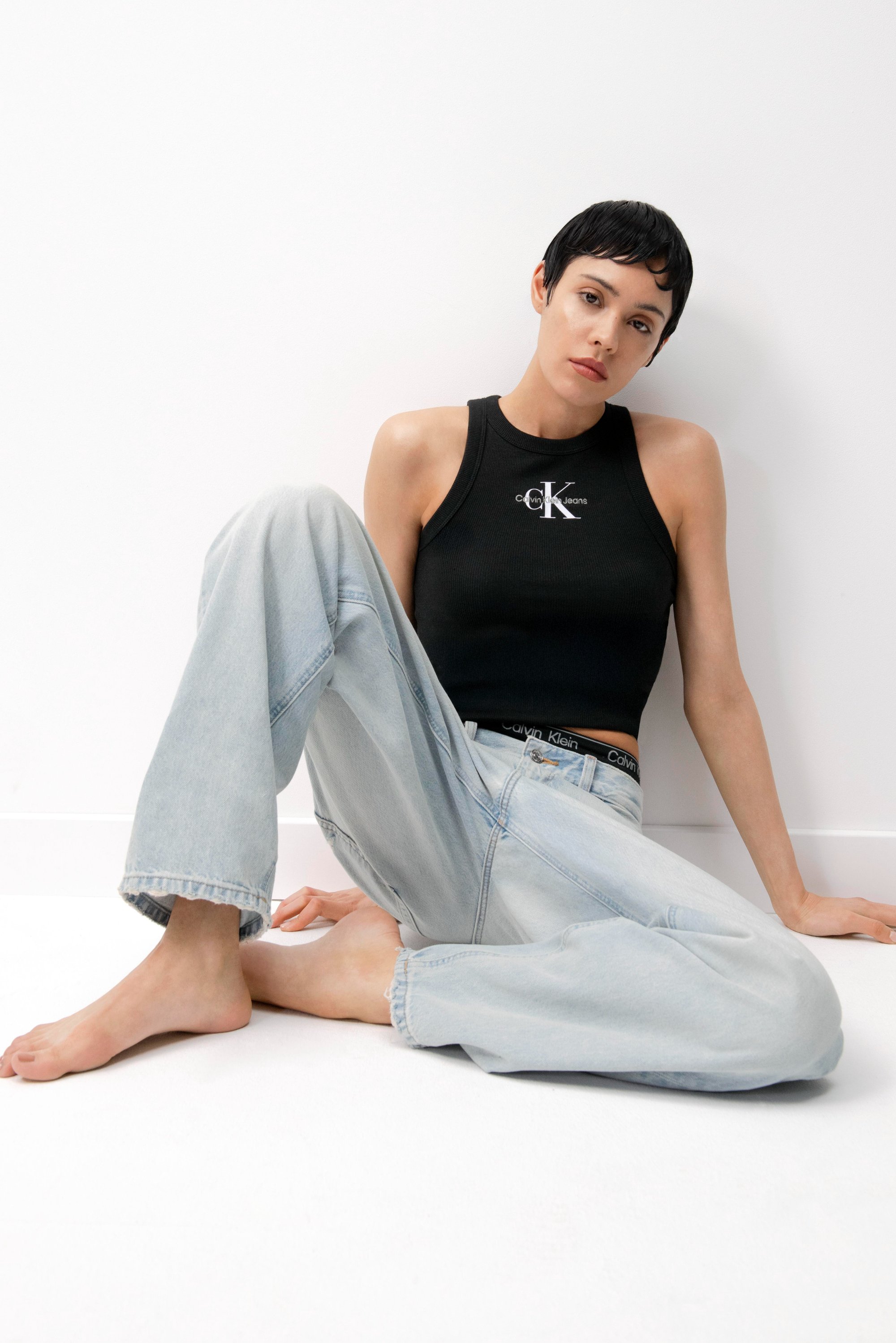 Een jonge vrouw met kort donker haar zit op de vloer, gekleed in een zwarte tanktop en een lichtblauwe baggy jeans, tegen een witte muur.