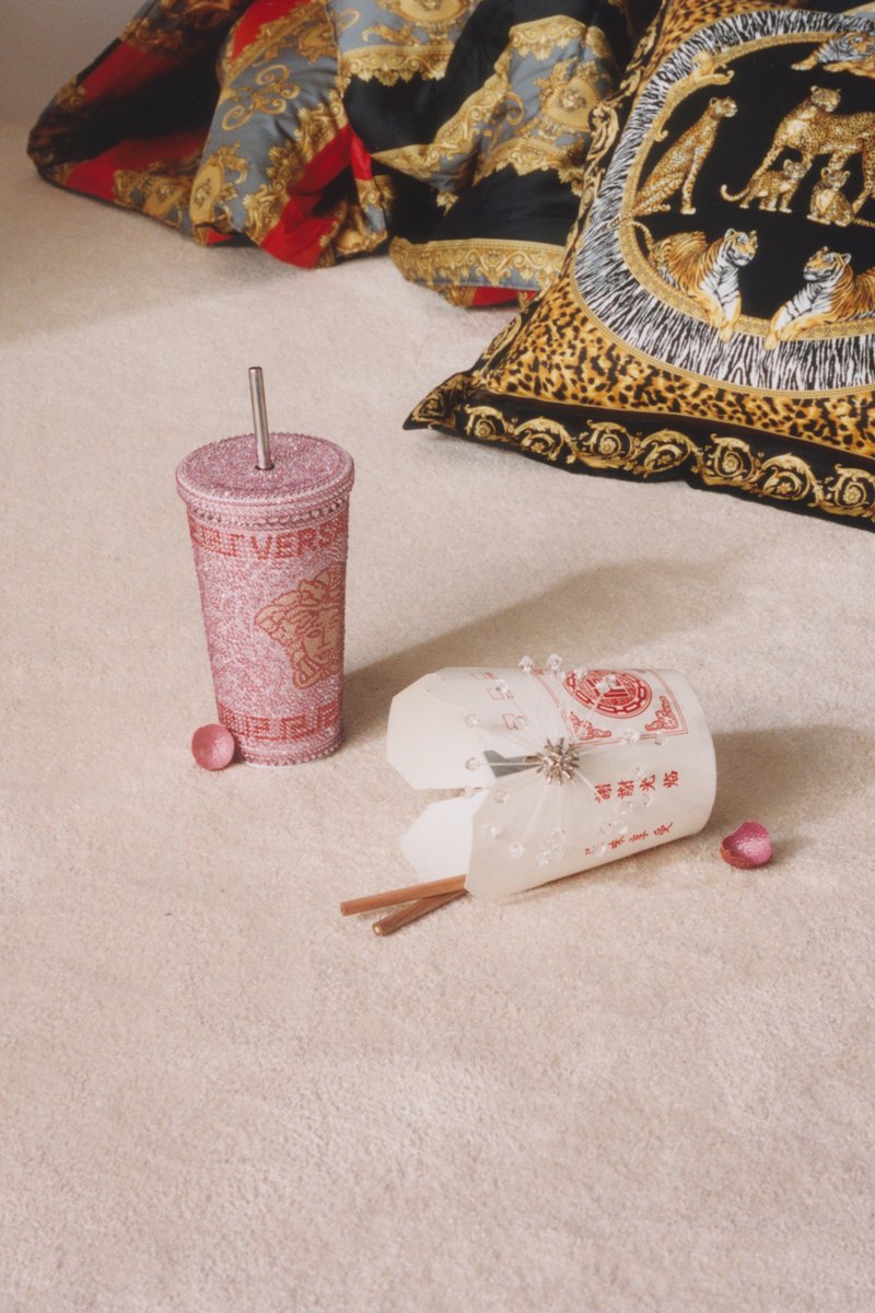 Rosa kopp med perler og sugerør samt hvit takeout-boks med spisepinner på beige teppe ved siden av utsmykkede puter i rødt, gull og leopardmønster.