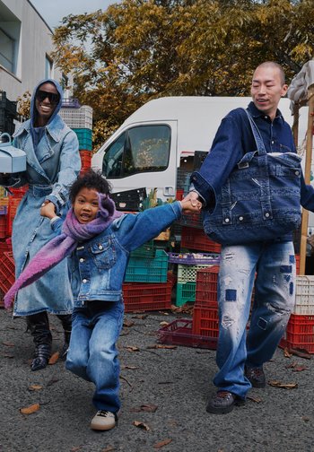 Three people outdoors in denim outfits: a long denim coat, a two-tone denim jacket with a purple scarf, and a large denim bag. Colorful crates in background.