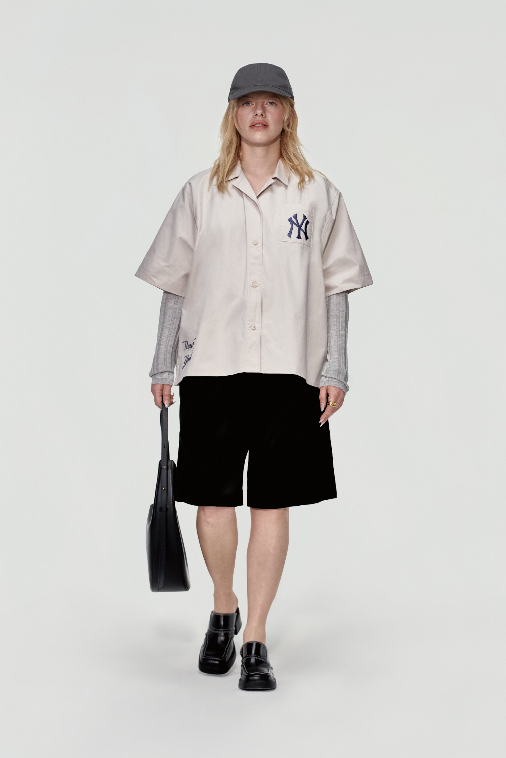 Una persona è in piedi contro uno sfondo semplice, indossando una camicia chiara con un logo, pantaloni corti neri e un cappellino, mentre tiene una borsa nera.