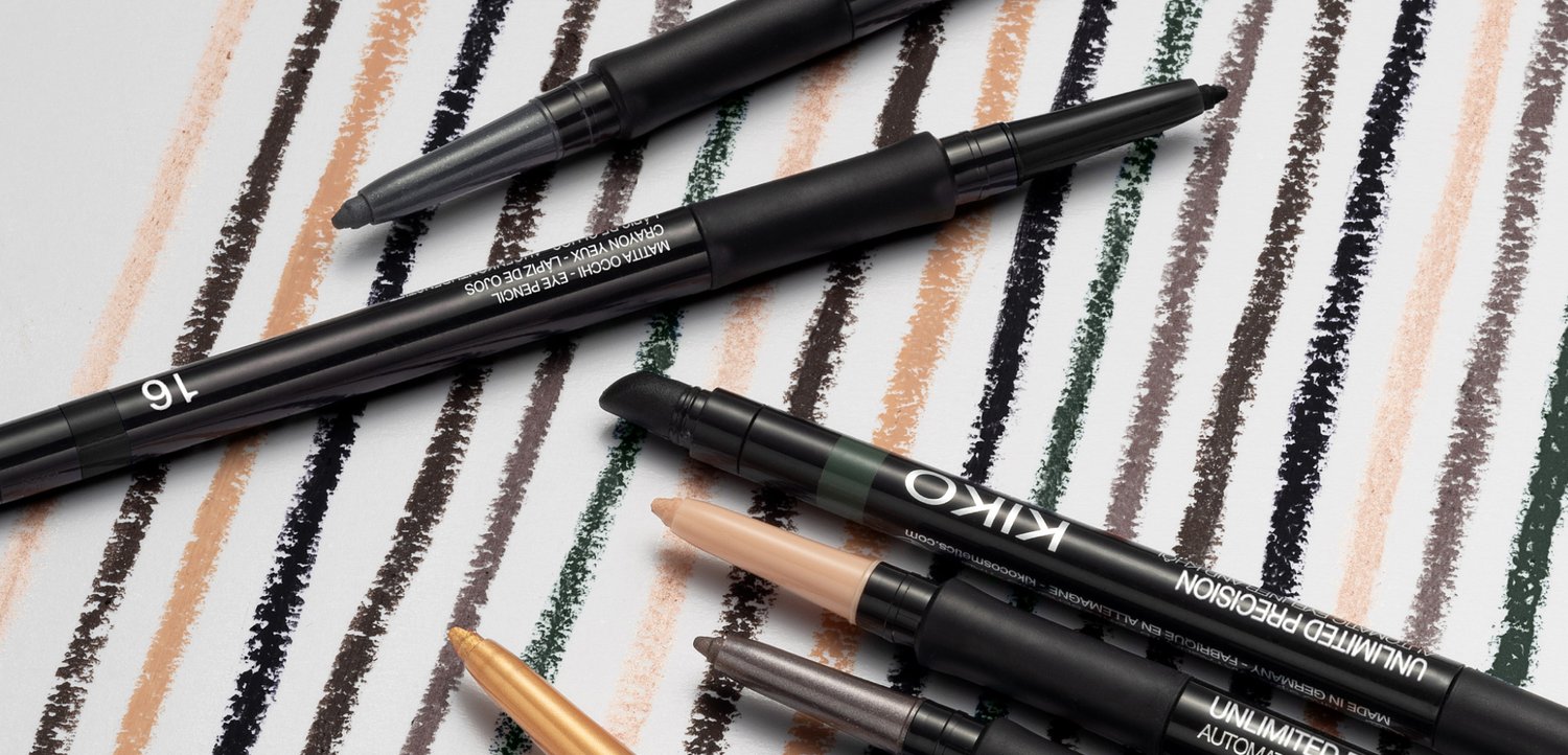 Plusieurs crayons de maquillage noirs et eyeliners reposent sur une surface avec plusieurs traits colorés ressemblant à des crayons de cire dans des nuances de brun, noir, vert et beige.