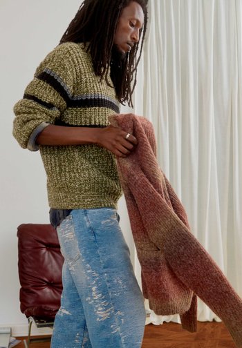 Hombre con rastas, vestido con un suéter verde y vaqueros rotos, sosteniendo un suéter marrón tejido, de pie en el interior cerca de una silla roja y cortinas blancas.