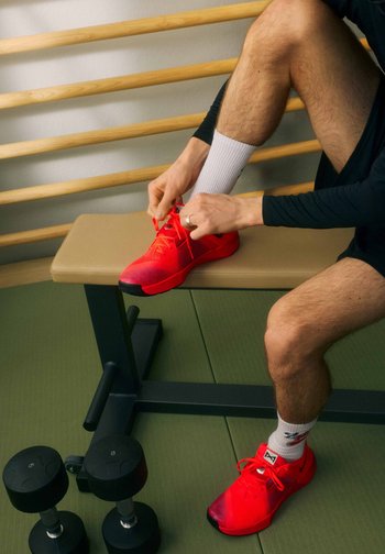 Person seated on bench ties bright red athletic shoes, wearing white socks and black athletic shorts near black dumbbells on green floor.