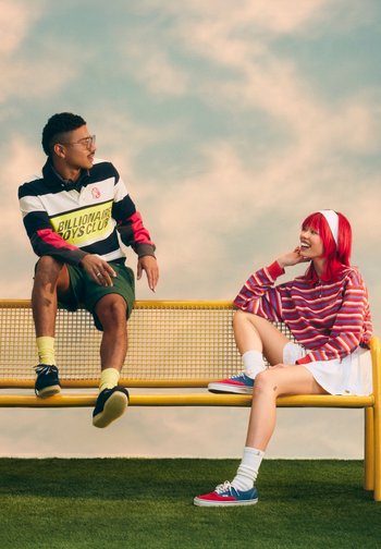 Dos jóvenes con ropa informal están sentados y hablan en un banco amarillo al aire libre bajo un cielo nublado.