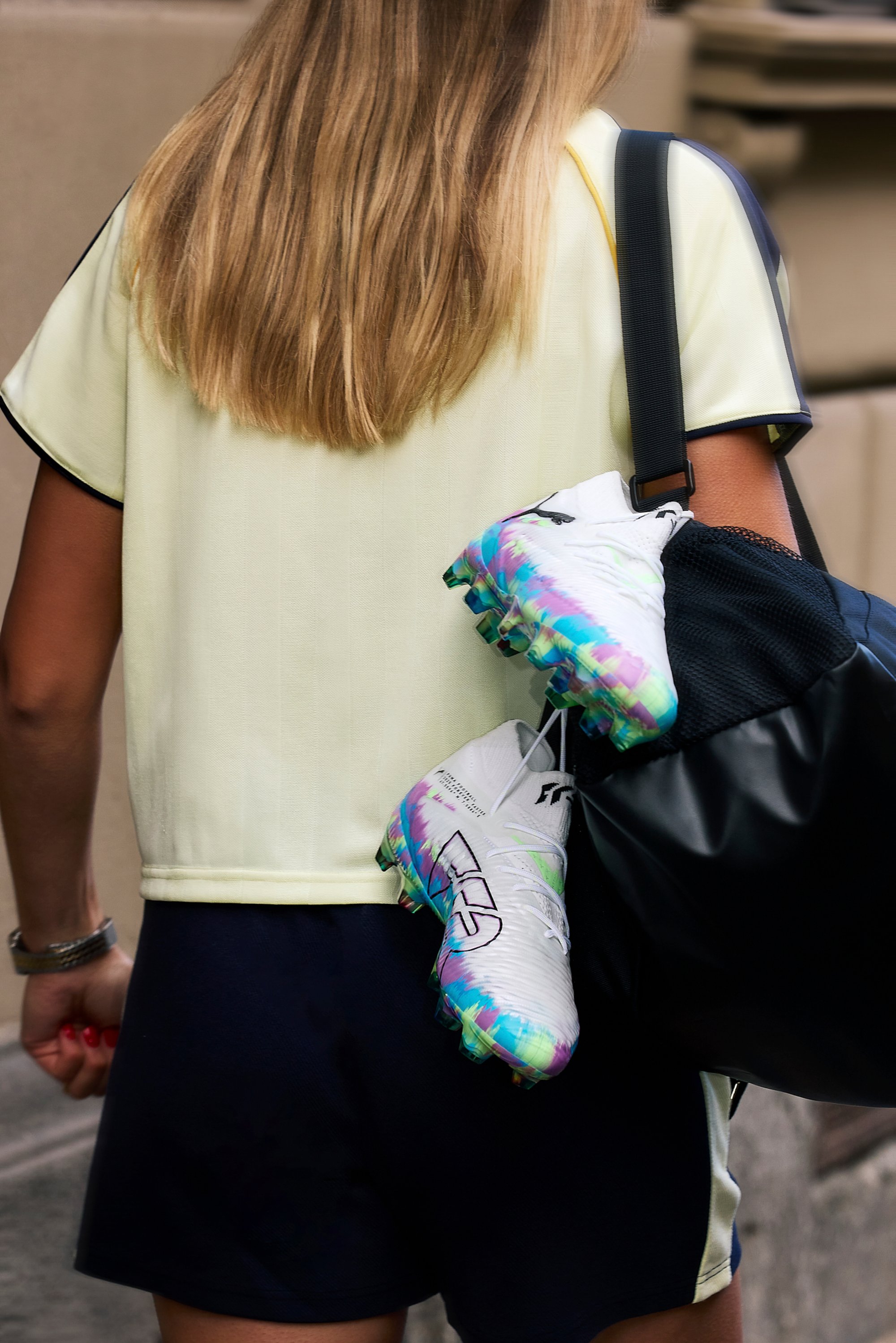 A person with long hair wears a yellow athletic top and navy shorts, carrying a black bag and colorful soccer cleats.