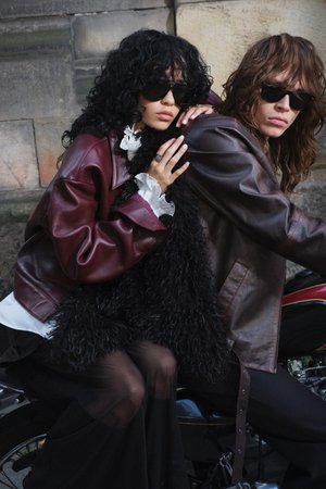 Two individuals are seated on a motorcycle, one wearing a maroon leather jacket and the other a brown leather jacket, both featuring textured black fur accents.
