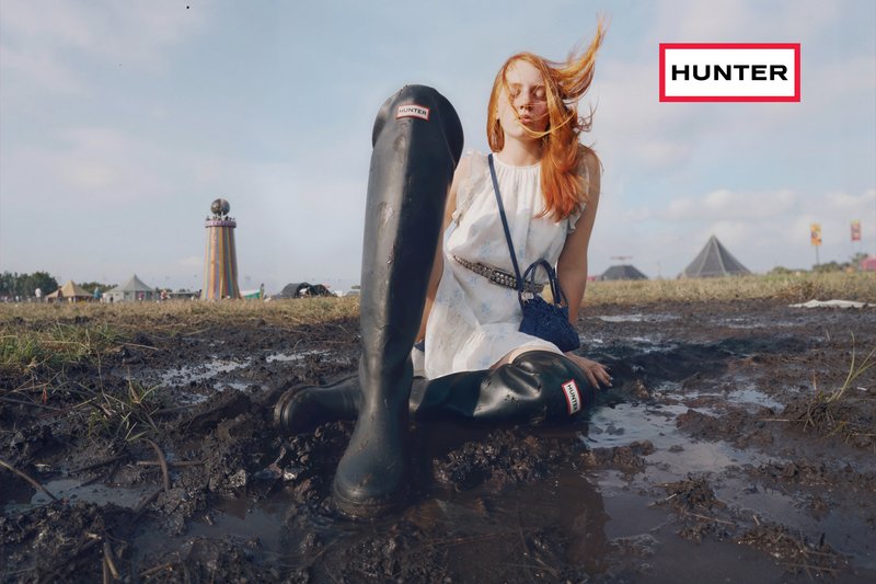 Mladá žena v bílých šatech sedí v bahnité vodě a má na sobě příliš velké černé boty Hunter na venkovním festivalu s stany a věží v pozadí.