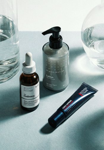 Prodotti per la cura della pelle su una superficie azzurro chiaro: flacone di siero The Ordinary, dispenser di lozione Skandinavisk, tubo di crema Biotherm Homme e due bicchieri d'acqua.