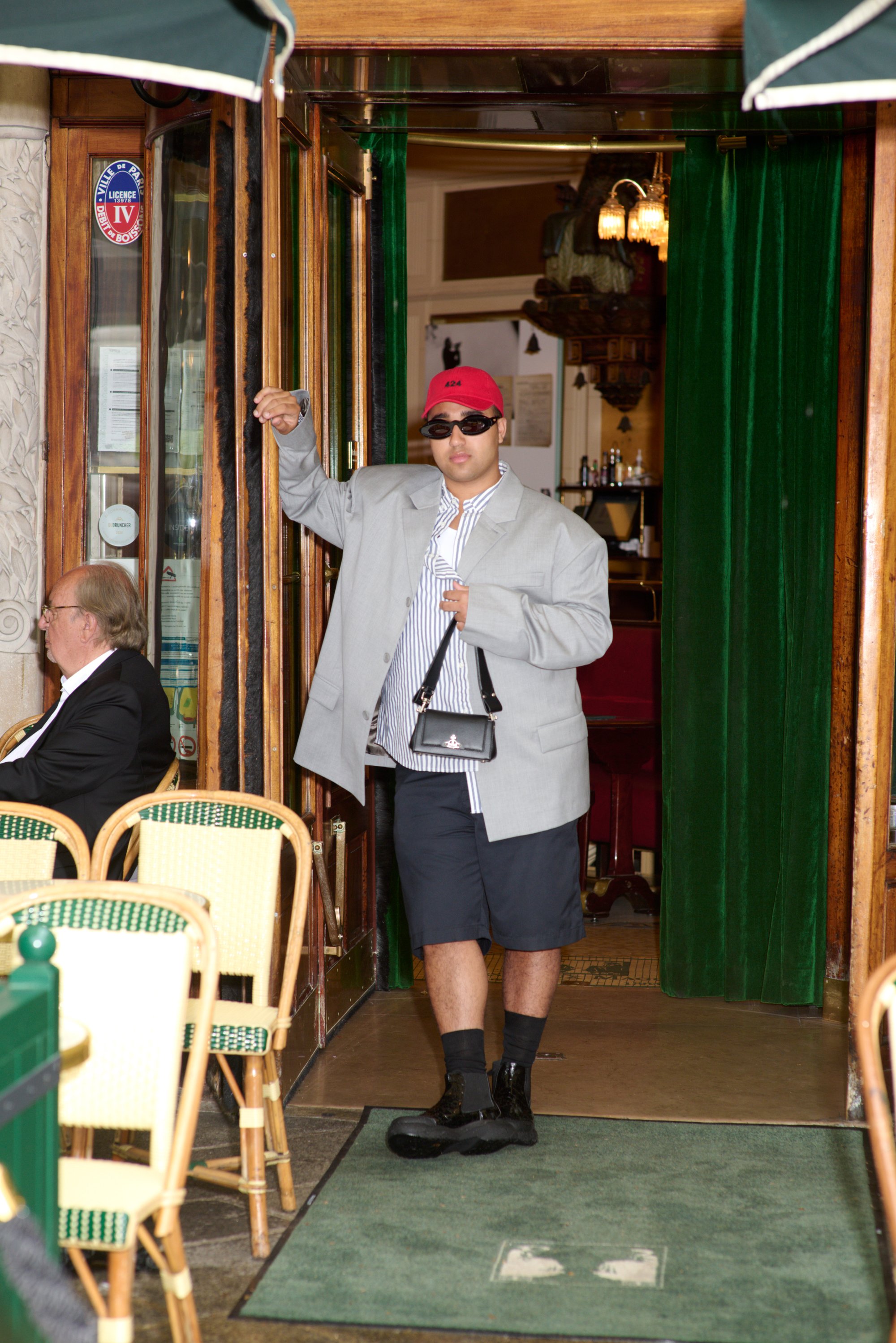 Una persona con un gran blazer gris, pantalones cortos negros y una gorra roja sostiene un bolso mientras sale de un café, con otro hombre sentado cerca.