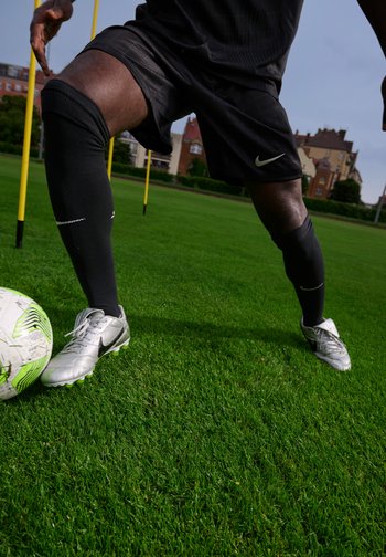 Crampons de football argentés sur l'herbe, associés à des chaussettes de compression noires et à un short, avec un ballon de football blanc et vert à proximité. Des cônes d'entraînement en arrière-plan.