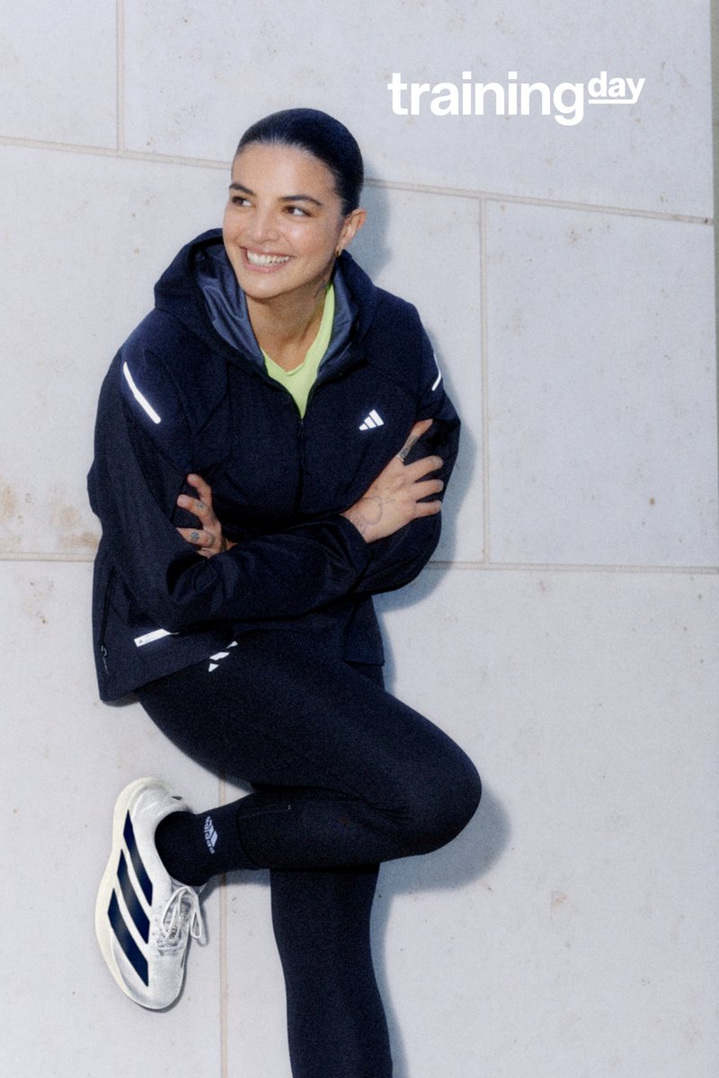 Mujer sonriendo con ropa deportiva, sentada contra una pared, con chaqueta negra y leggings, y zapatillas blancas y azules. Texto "día de entrenamiento" en la esquina.