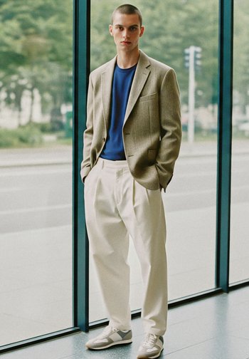 Completo da uomo con un blazer beige chiaro, camicia blu, pantaloni bianchi plissettati e sneakers grigie. Il blazer ha un colletto a rever e tasche.