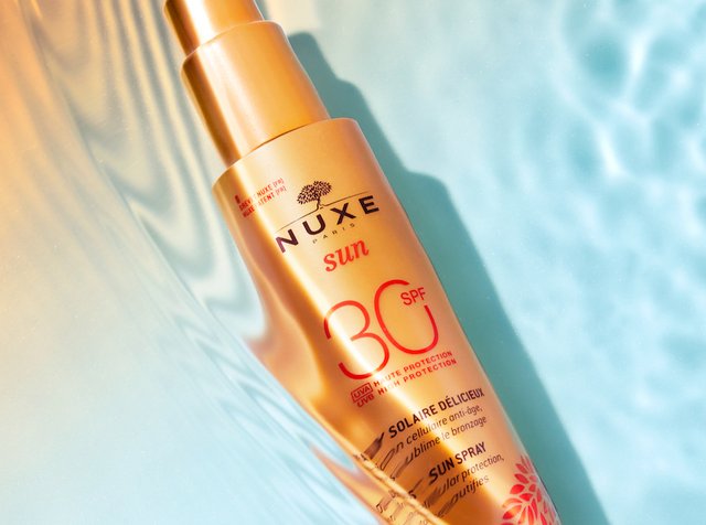 Botella de spray dorada etiquetada como "NUXE sun SPF 30" con texto que resalta las características de protección solar, descansando sobre un suave fondo azul.