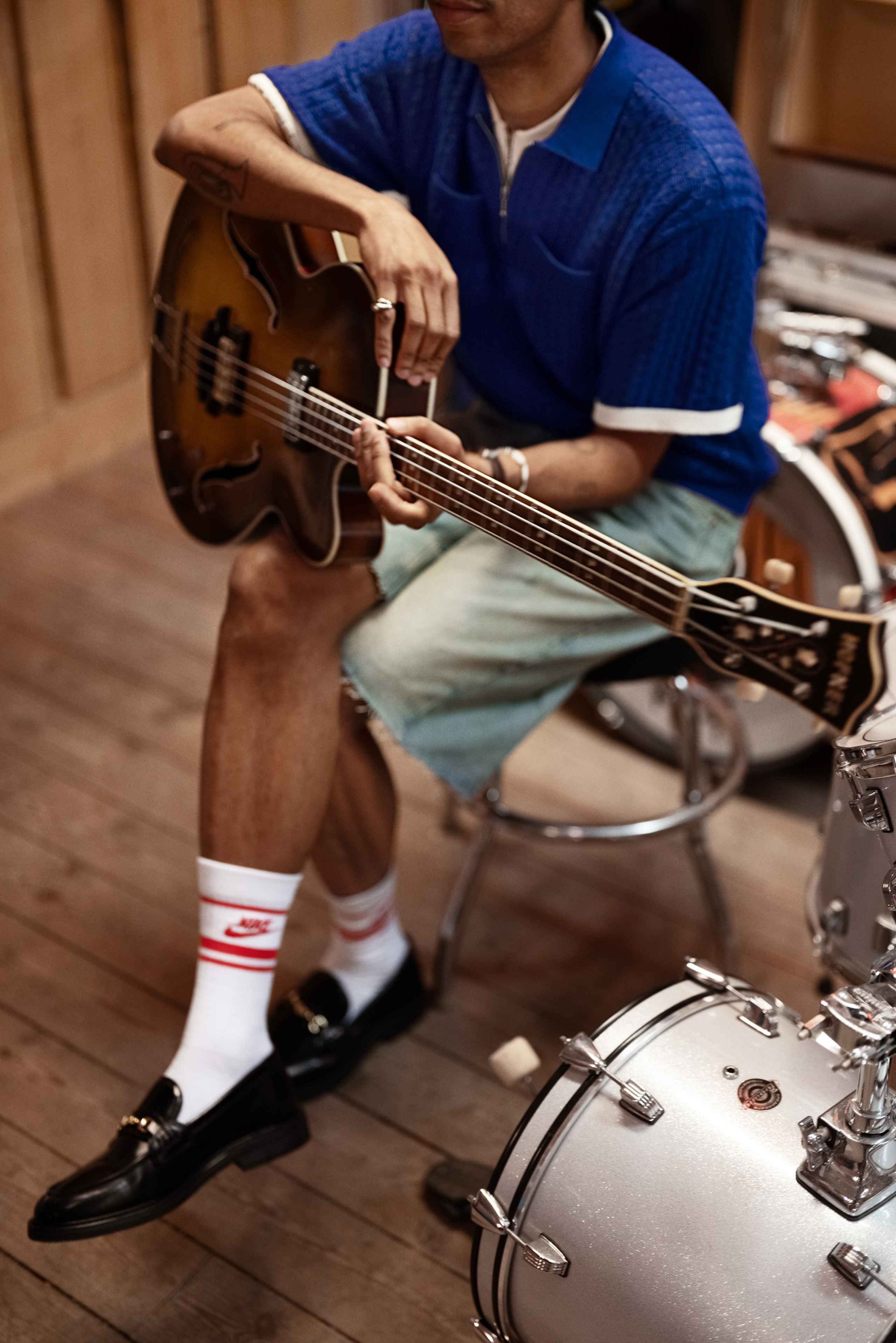 Bruine elektrische basgitaar vastgehouden door een persoon in een blauw polo met korte mouwen, denim shorts, zwarte instappers en witte sokken met rode strepen.