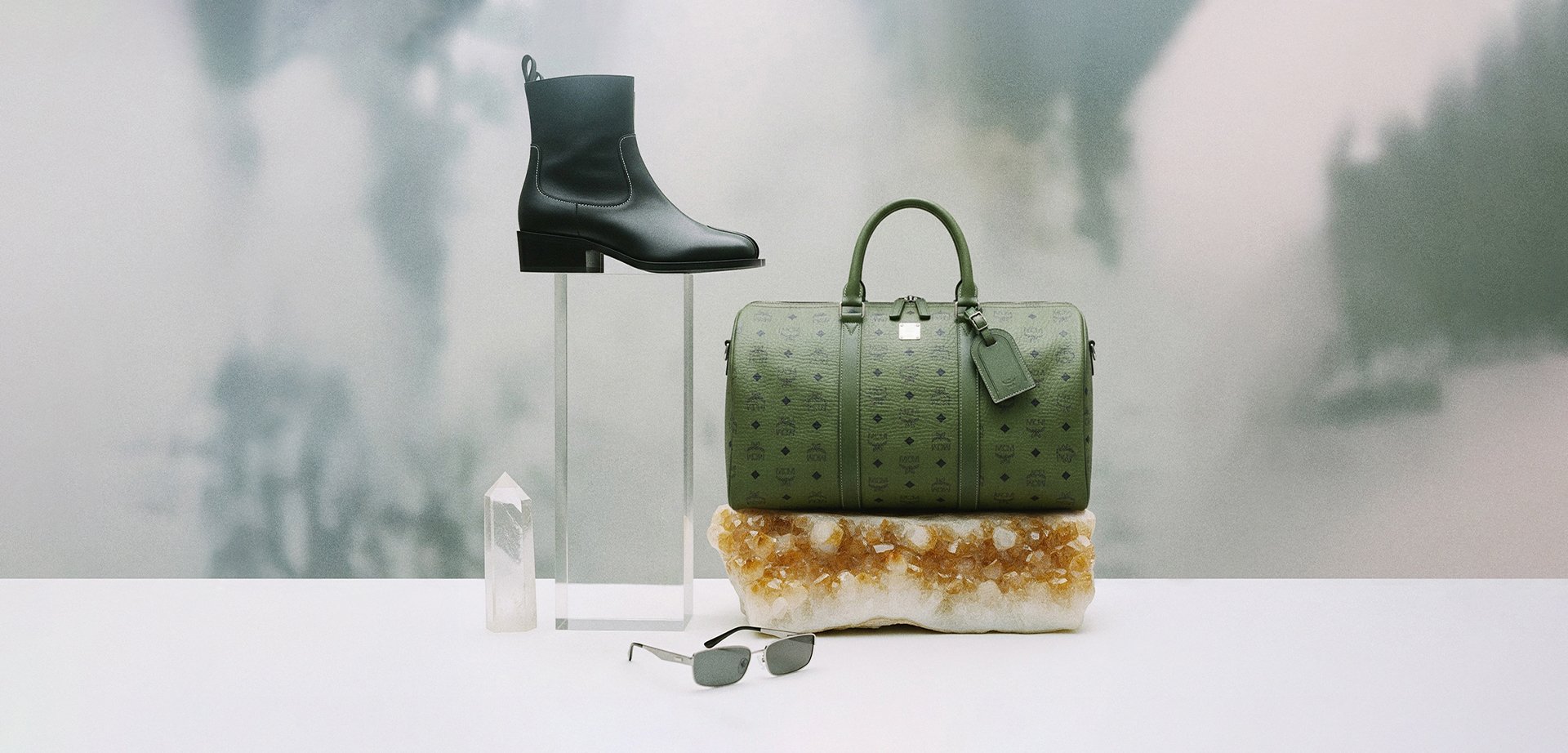 Negro bota de tobillo de cuero, bolso de viaje de cuero texturizado verde, acento de cristal y gafas de sol rectangulares sobre un fondo claro con un sutil degradado.