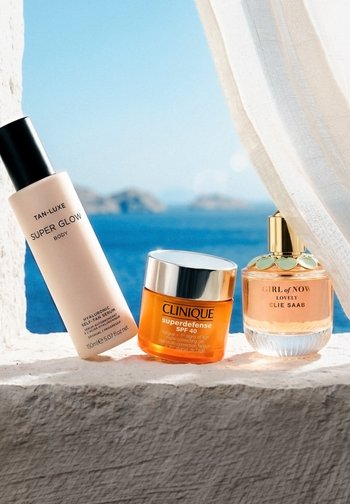 Sérum corps Tan-Luxe Super Glow, gel Clinique Superdefense SPF 40 et parfum Elie Saab Girl of Now sur un rebord blanc au bord de la mer bleue sous un rideau léger.