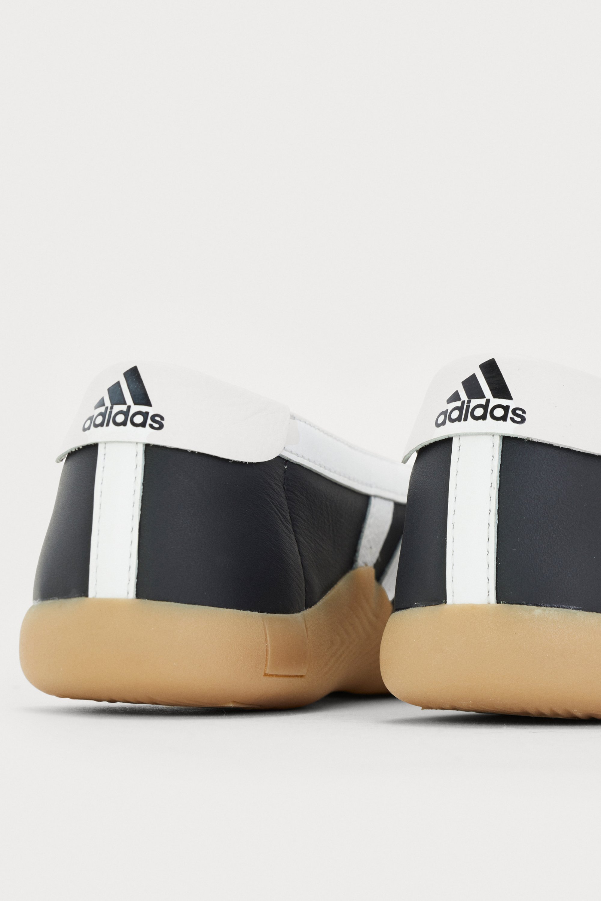 Black and white Adidas shoes displayed from the rear, showcasing a white logo on the heel and a gum sole, set against a plain background.