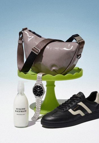 Sac bandoulière taupe sur un présentoir à gâteau vert, montre métal argentée suspendue au présentoir, bouteille blanche de lotion pour le corps et basket noire en cuir sur surface blanche.