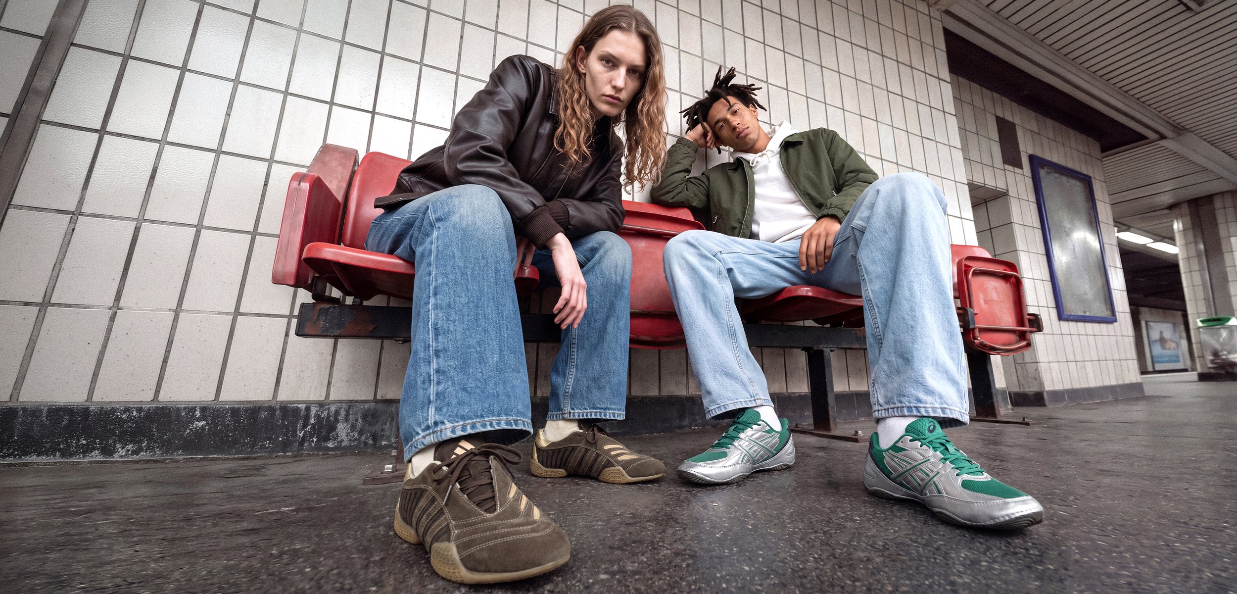 Deux jeunes hommes en tenue décontractée sont assis sur un banc rouge dans une station de métro carrelée et regardent vers l’appareil photo.