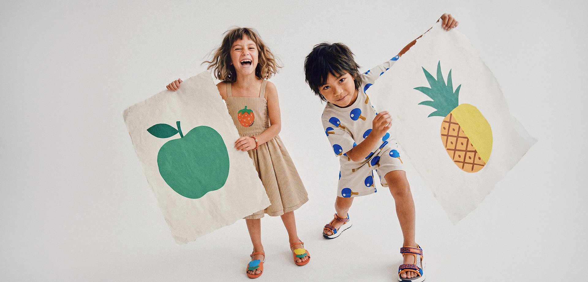 Zwei Kinder halten große Fruchtzeichnungen, eines mit einem grünen Apfel und das andere mit einer gelben Ananas, beide lächeln und sind verspielt.