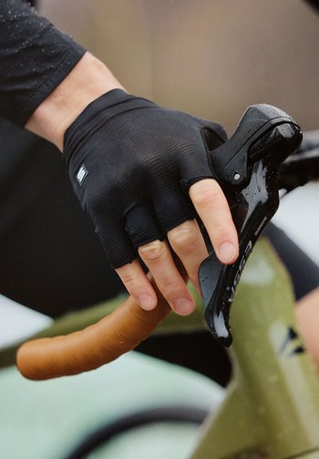 Hånd iført svart fingreløs hanske som holder et regndekket sykkelbremsespake på en sykkel med brunt styreband.