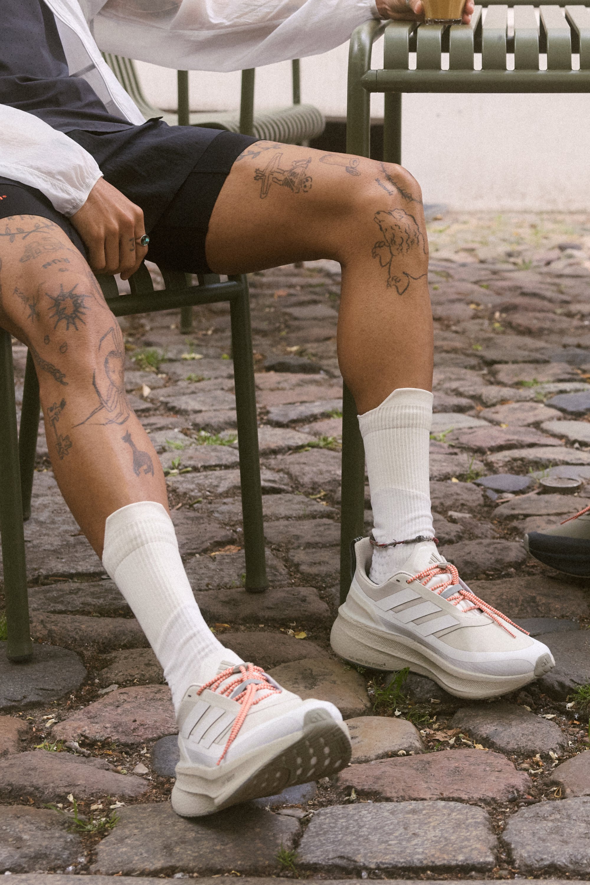 Una persona è seduta su una sedia verde, indossando pantaloni corti neri, calze bianche e scarpe bianche con lacci rosa, mostrando vari tatuaggi sulle gambe.