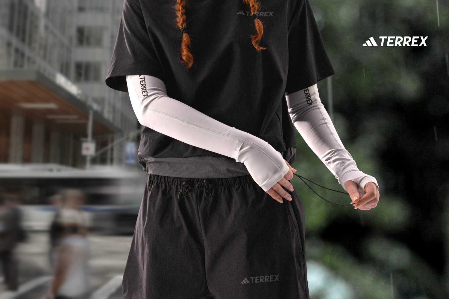 Persona con cabello trenzado rojo ajusta el cordón de unos pantalones cortos negros, viste una camiseta negra y mangas de protección UV blancas. Fondo urbano.