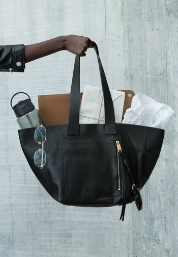 Main tenant un sac fourre-tout en cuir noir contenant des lunettes de soleil, une bouteille d'eau, un dossier marron et des papiers, devant un mur en béton gris.