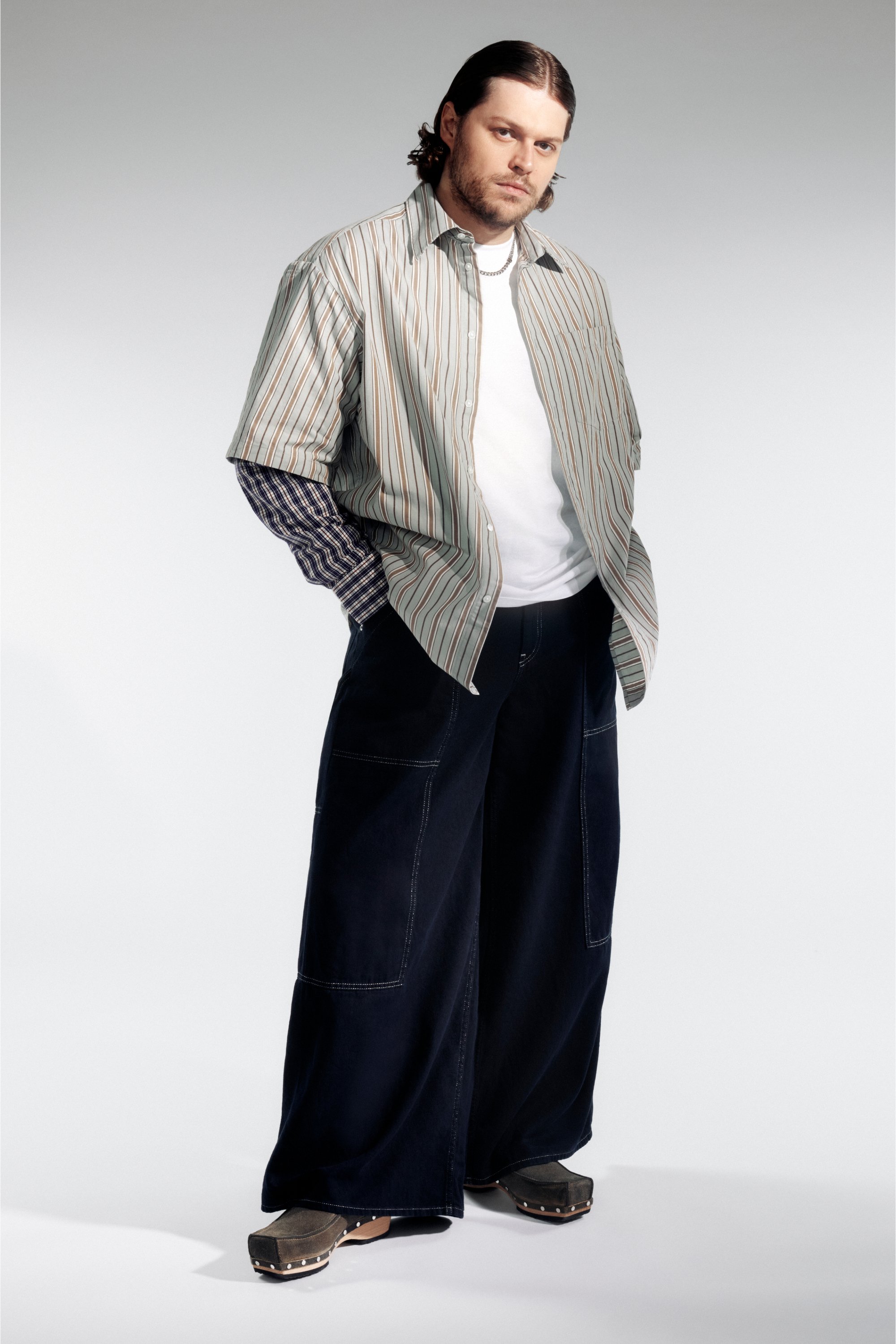 Un hombre se encuentra de pie con confianza, vistiendo una camisa de rayas de gran tamaño sobre una camiseta blanca, pantalones oscuros de pierna ancha y zapatos únicos, con un fondo neutro.